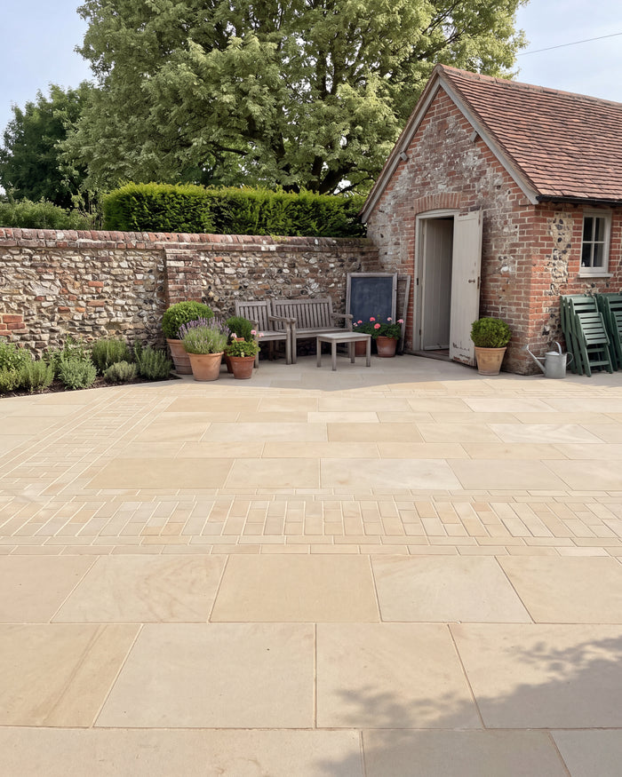 Wensley York Sandstone Honed paving across a walled courtyard beside a brick and flint outbuilding, with a weathered teak bench and terracotta pots of lavender